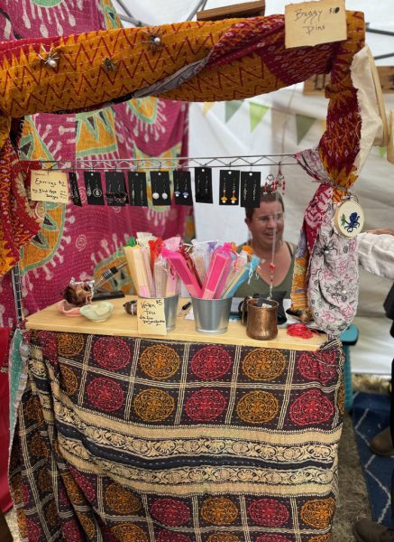 Photo showing me sitting behind my faire booth table, smiling, with many colorful blankets draped for decoration.